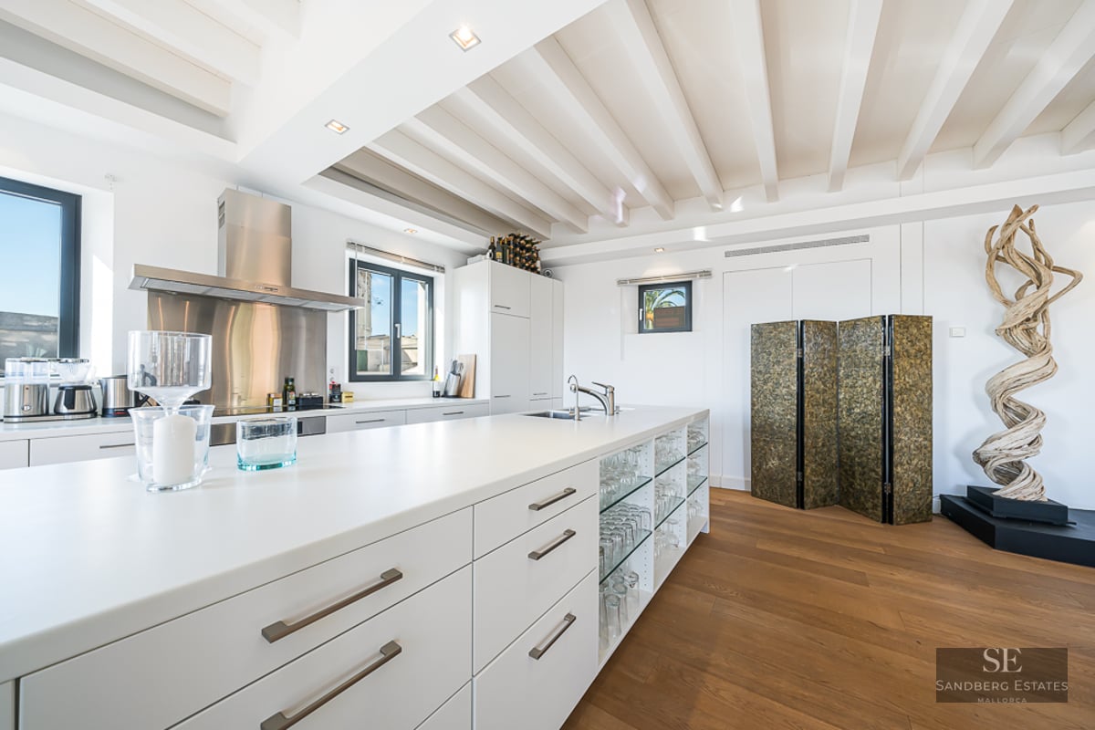 Modern white kitchen featuring a large island, wood flooring, ceiling beams, and a decorative wood sculpture.