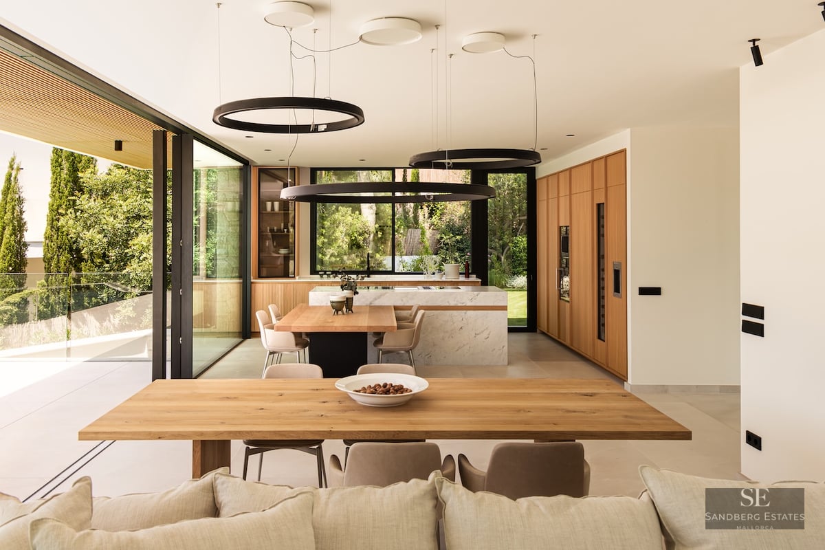 High-end kitchen with a white marble island, wooden cabinets, and a long dining table under circular halo pendant lights.