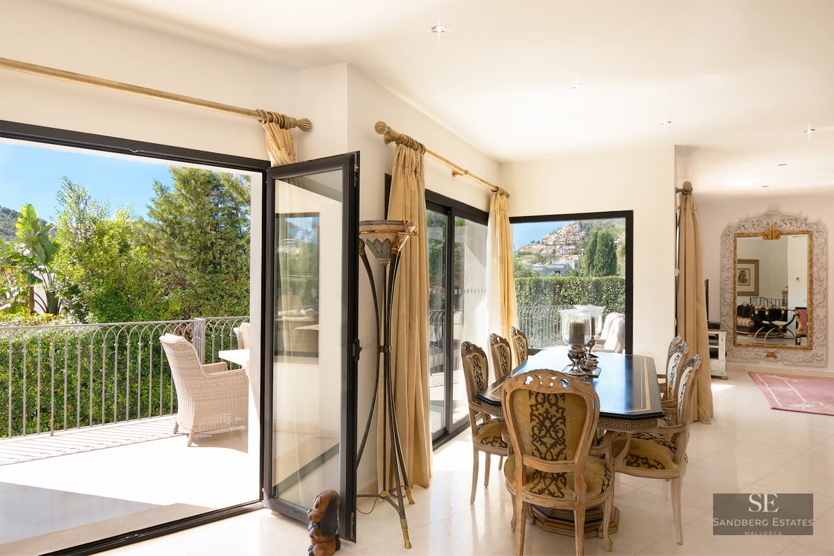 Luxury dining room featuring a black table, ornate chairs, and large open glass doors leading to a balcony with lush greenery.