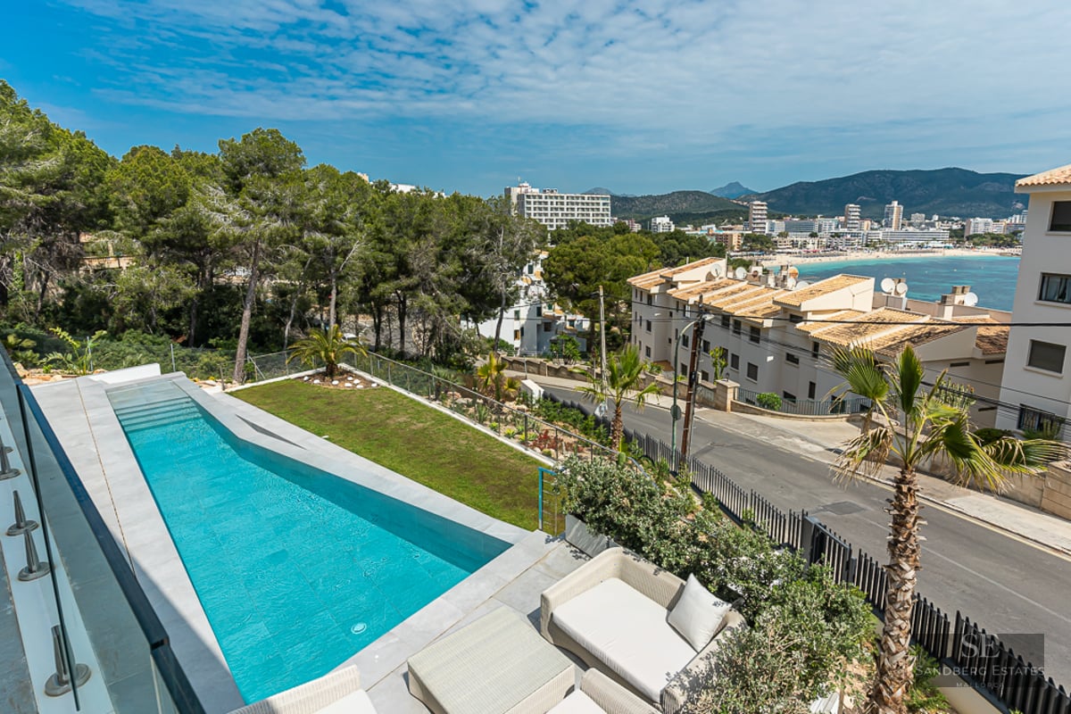 A long turquoise swimming pool on a terrace with glass railings, overlooking a coastal town and distant mountains.