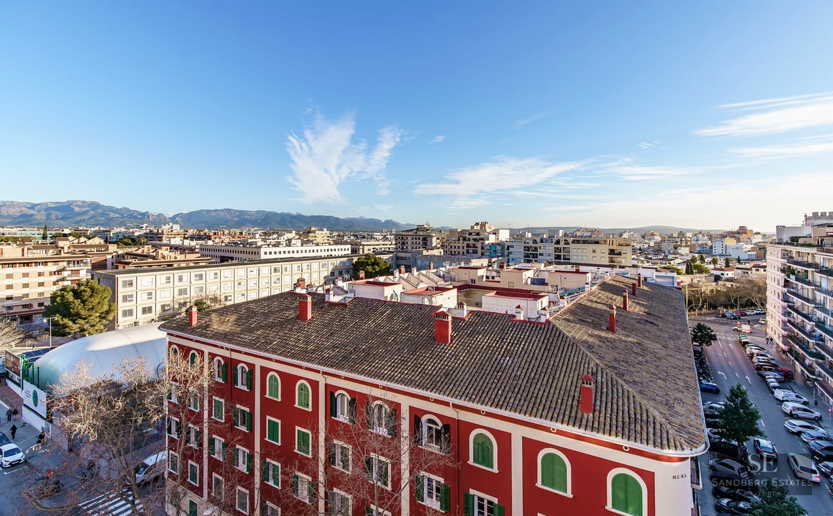 High-angle city view featuring a red building with tiled roofs, urban streets, and distant mountains under a clear sky.
