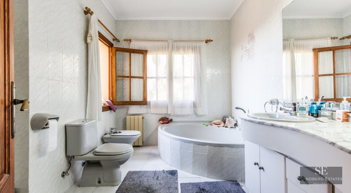 Spacious bathroom featuring a large corner bathtub, marble-topped vanity, toilet, bidet, and wooden-framed windows.