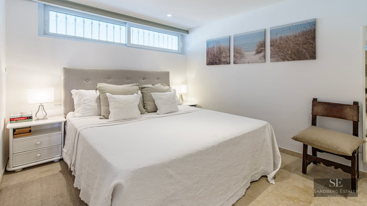 A bright bedroom featuring a large bed with a tufted headboard, white bedding, and coastal artwork on the wall.
