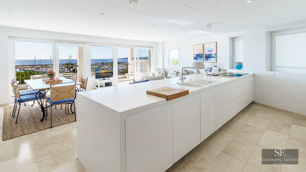 Minimalist white kitchen island leading to a dining area with a panoramic view of the harbor and sea.