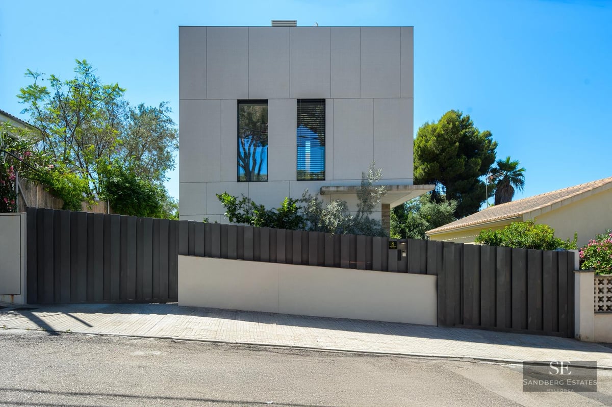 Exterior view of a modern white cubic house with a dark grey metal fence and green trees under a clear blue sky.