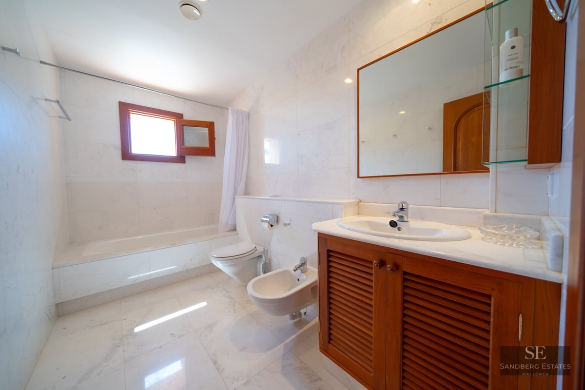 Bathroom featuring white marble walls and floors, a wooden vanity, toilet, bidet, and bathtub under a window.