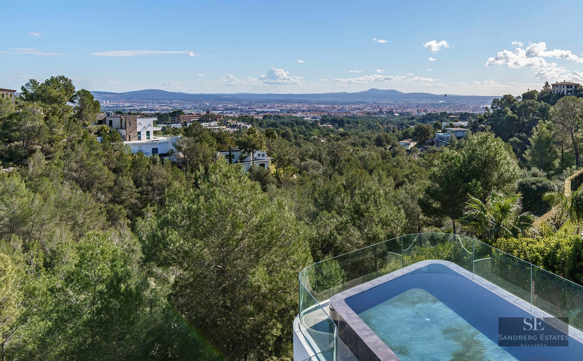 An infinity pool with a glass railing overlooking a lush green valley, a sprawling city, and distant mountains.
