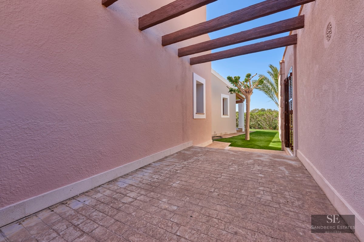 A stone-paved outdoor passage with pink stucco walls and dark wooden beams leading to a green garden.