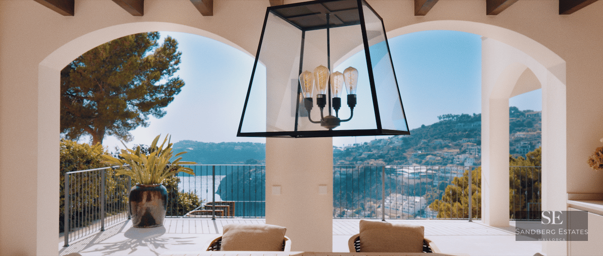View through white arches from a villa terrace overlooking the sea and green hills under a bright blue sky.