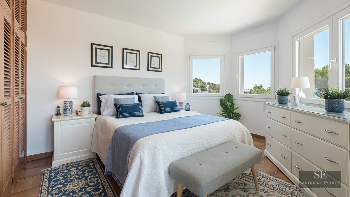 Bright bedroom with a white bed, blue pillows, white dresser, large windows, and terracotta tile floors.