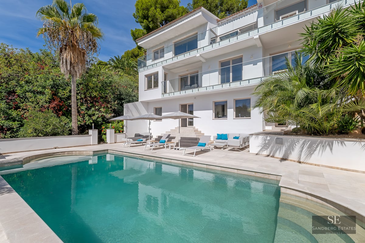 Three-story white villa featuring a large turquoise pool, sun loungers, and palm trees under a clear blue sky.