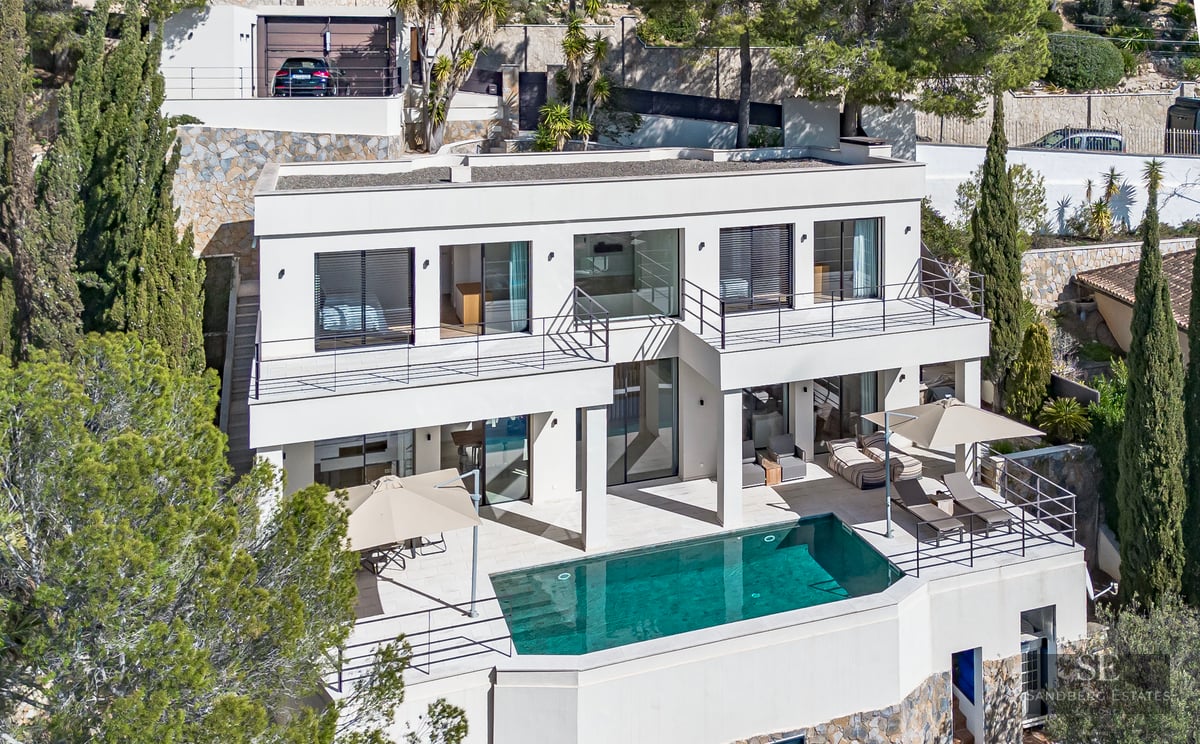 Aerial view of a white modern villa with a turquoise pool, sun loungers, and pine trees on a hillside.