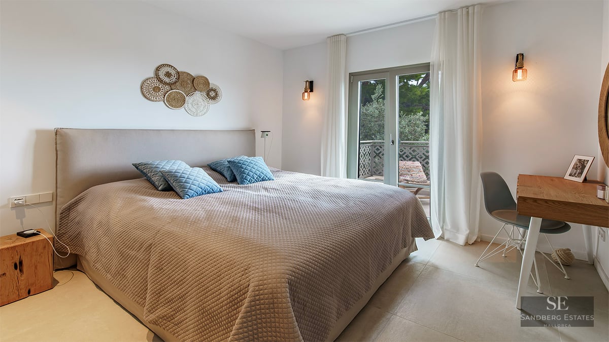 Modern bedroom featuring a large bed, beige textured bedding, blue pillows, wooden desk, and glass door to a balcony.