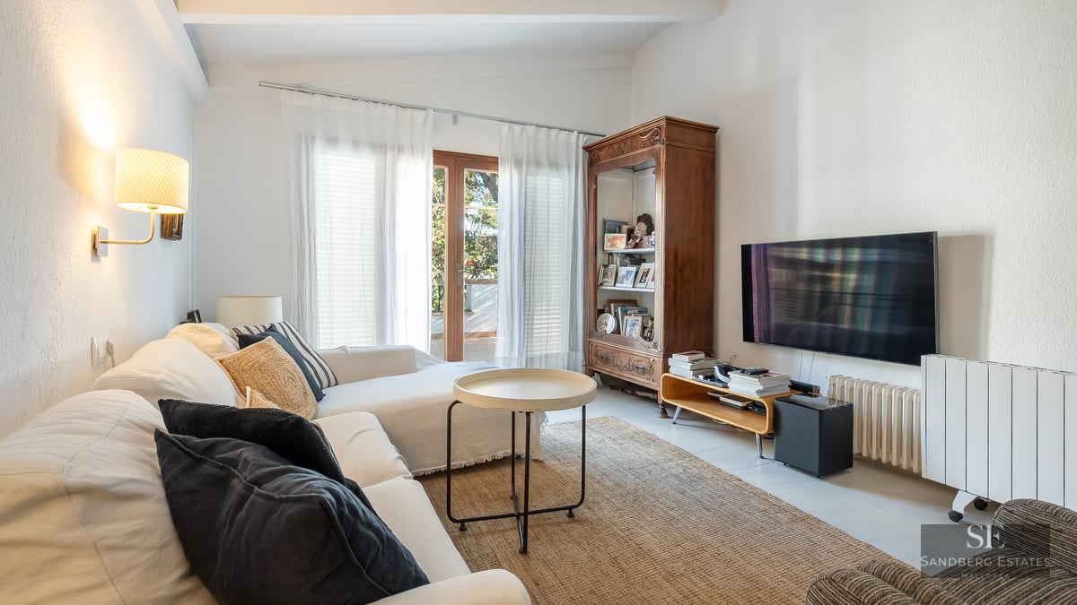 Bright living room with white sofas, a large wooden armoire, wall-mounted TV, and natural light from a terrace door.
