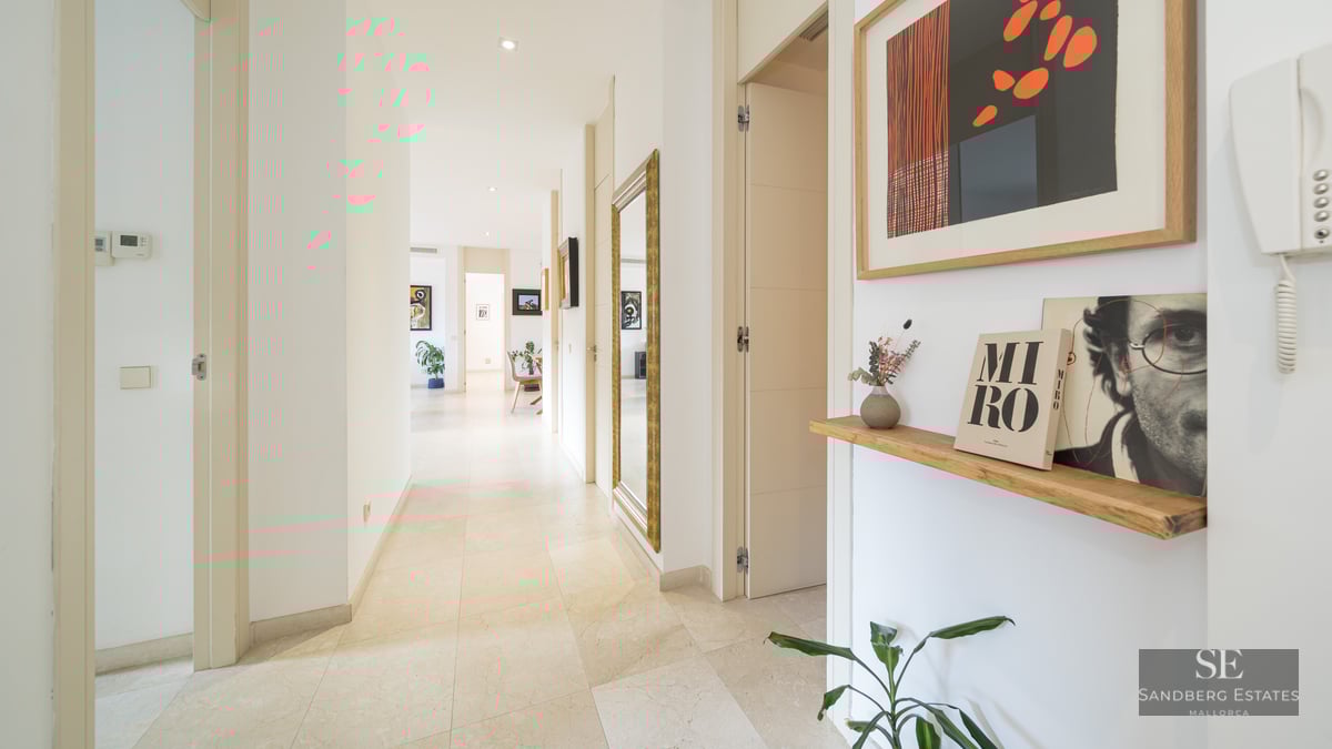 Long white hallway with stone floors, a large gold-framed mirror, and colorful art pieces.