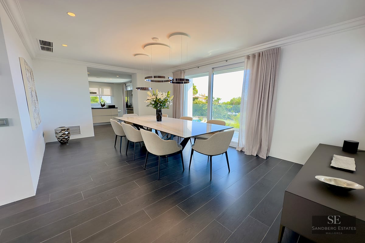 Minimalist dining room featuring a marble table, six beige chairs, dark flooring, and large windows viewing a garden.