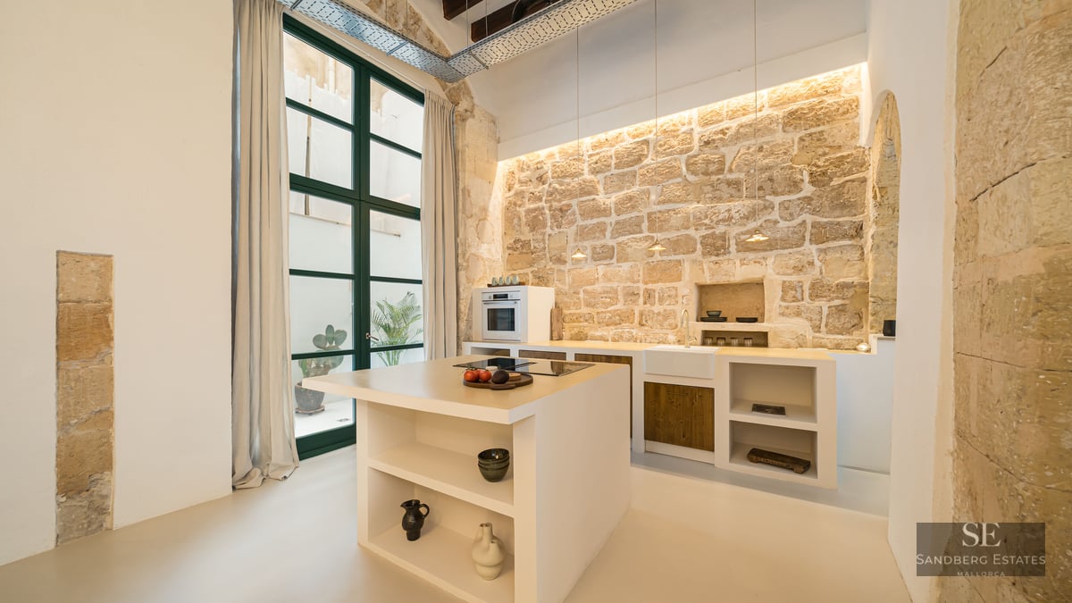 Cuisine moderne avec îlot blanc, mur en pierre rustique, hauts plafonds et grandes fenêtres.