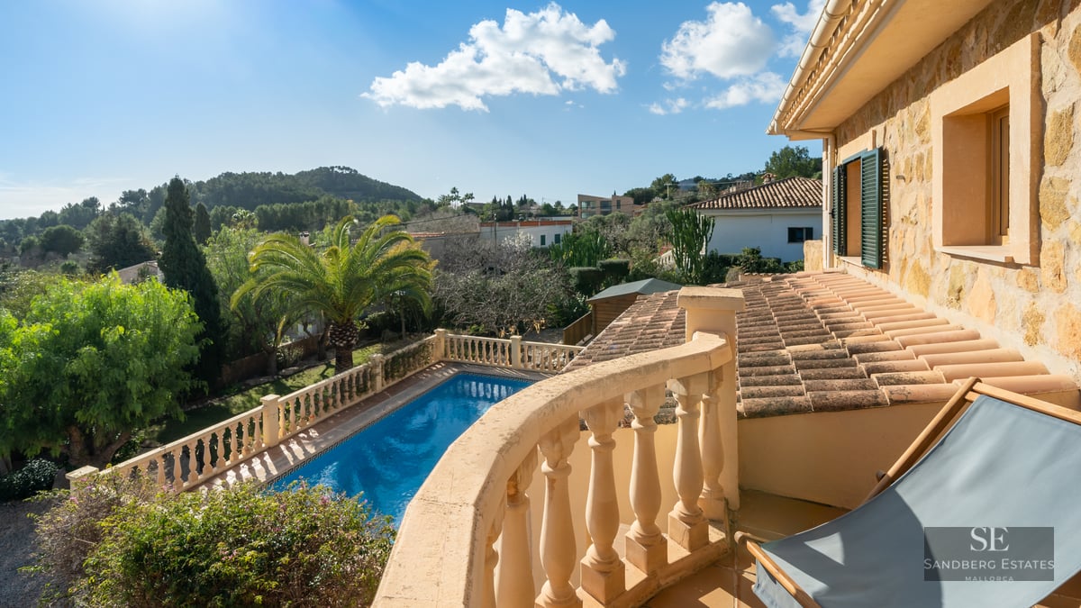 View from a stone balcony overlooking a blue swimming pool, lush gardens, and rolling hills under a clear blue sky.