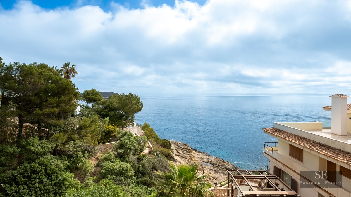 Elevated view of a rocky coastline with deep blue sea, green pine trees, and a Mediterranean villa.