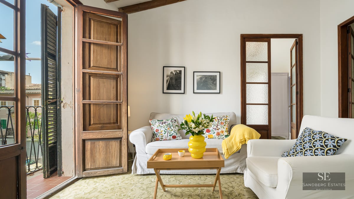 Bright living room with white sofas, colorful cushions, and large wooden balcony doors opening to a city view.