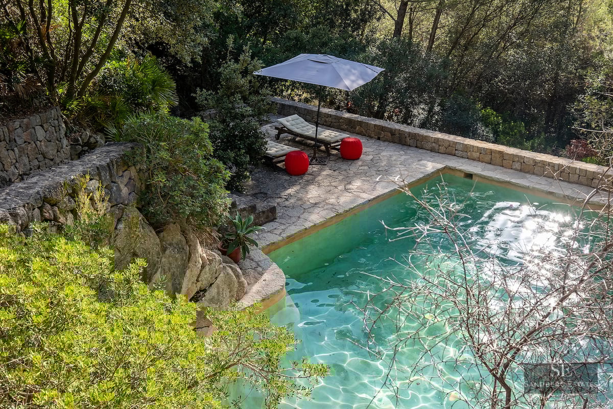 Elevated view of a turquoise swimming pool with stone deck, sun loungers, and red poufs nestled in a dense forest.