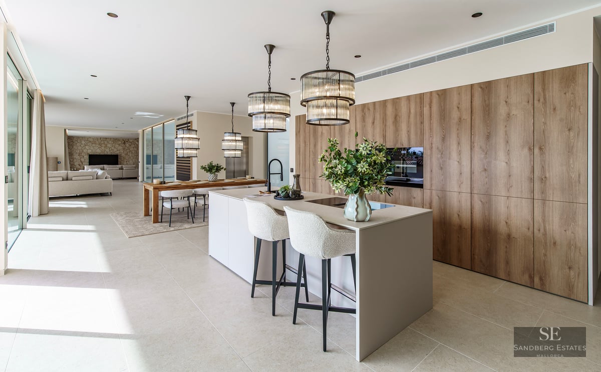 Modern kitchen with a large island, white stools, designer pendant lights, and wood cabinetry in an open-plan space.