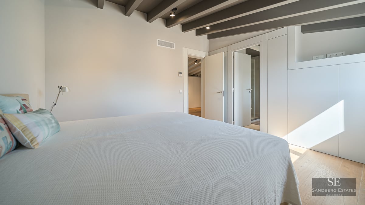 Bright bedroom featuring exposed ceiling beams, white bedding, built-in wardrobes, and light wood flooring.