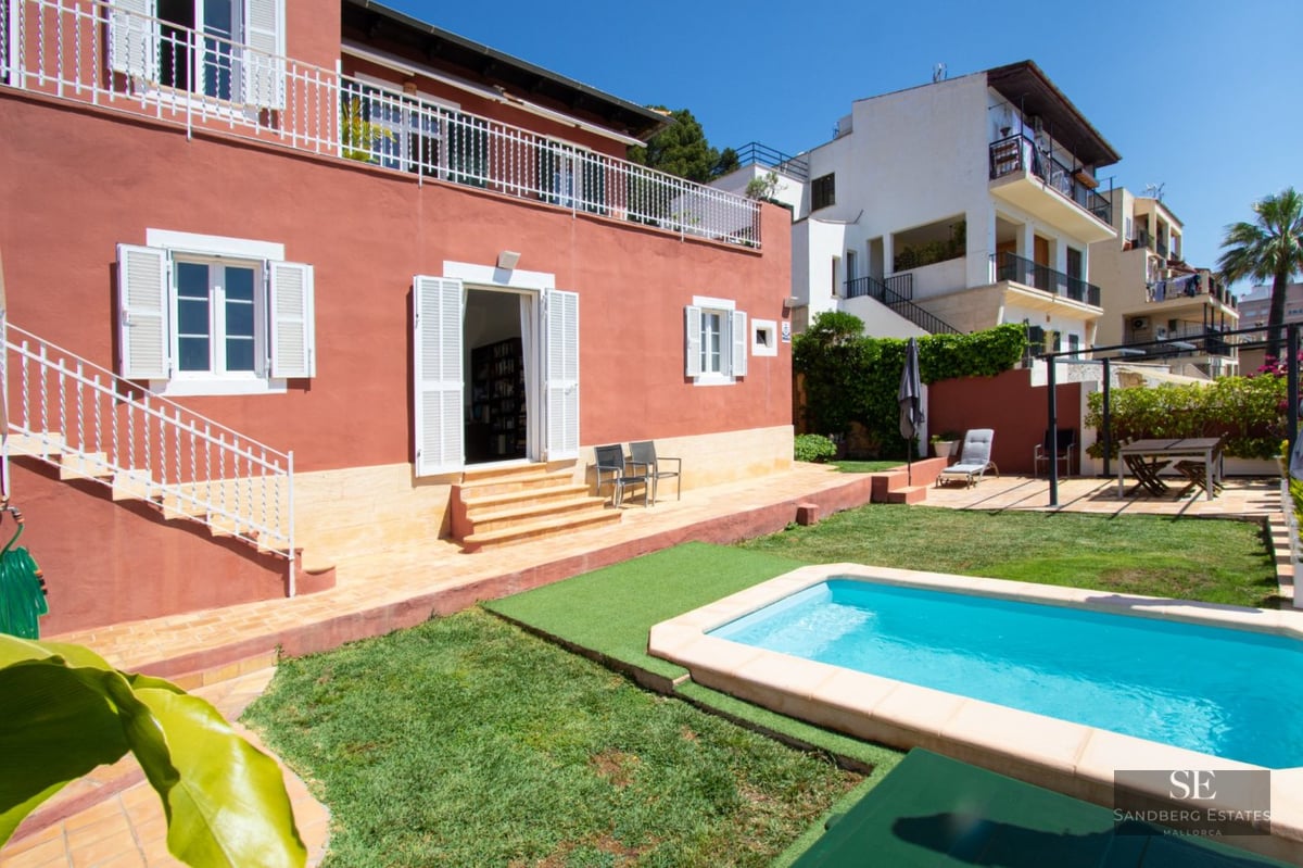 Terracotta-colored villa with a bright blue pool, lawn, and white shutters under a clear sky.