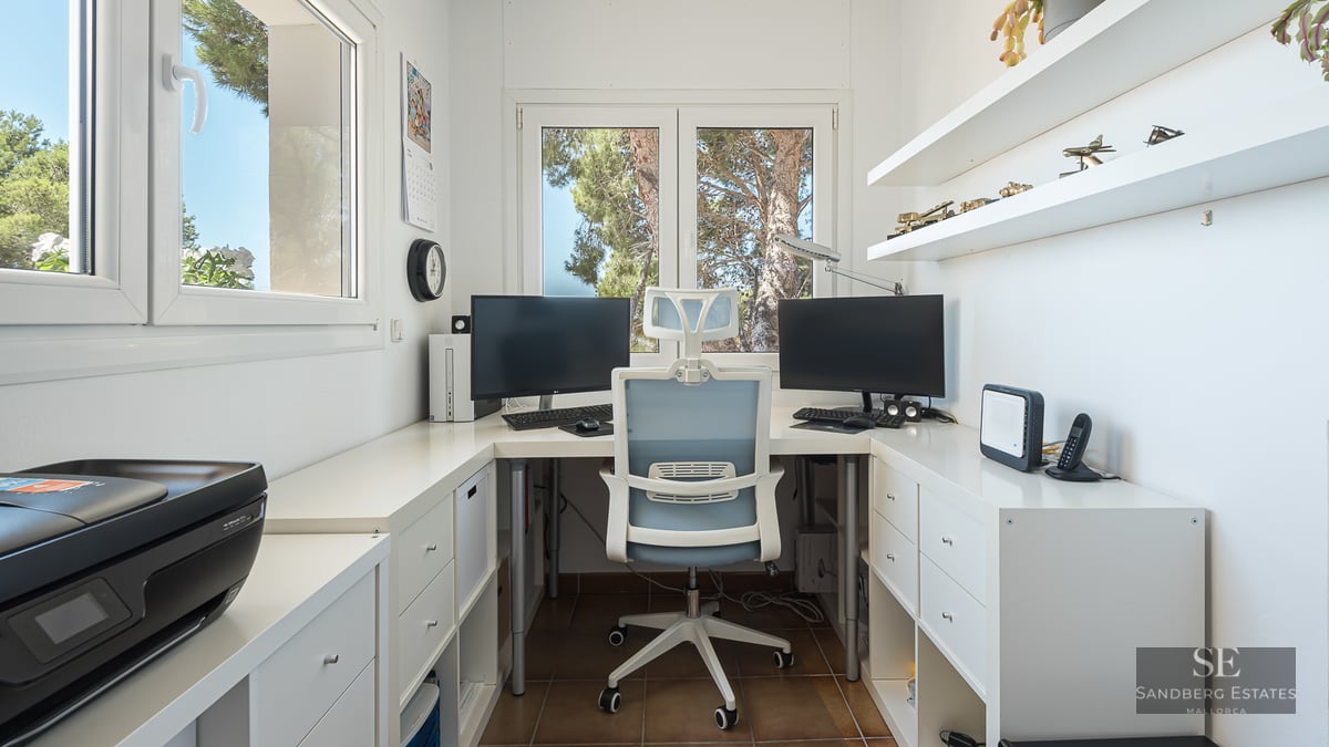 Modern white desk with dual monitors, ergonomic chair, and windows overlooking green trees.