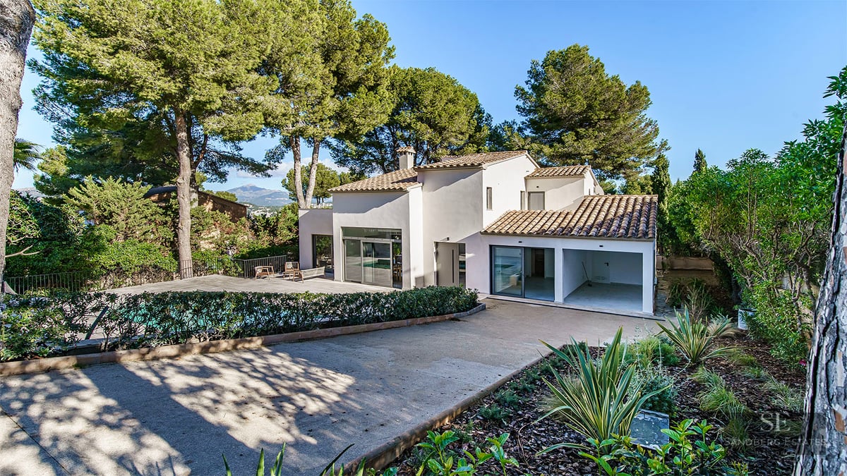 A two-story modern white villa with terracotta roof tiles, surrounded by tall pine trees and a swimming pool area.