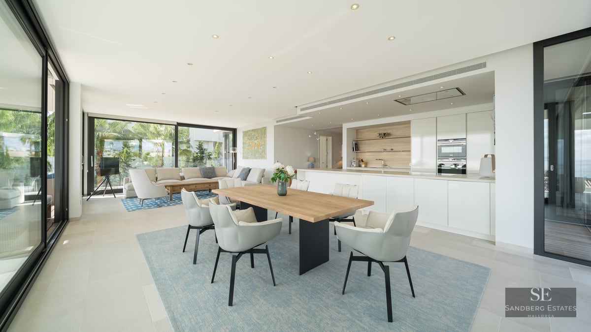 Bright open-plan room with a wooden dining table, white kitchen, and sofas next to large glass doors.