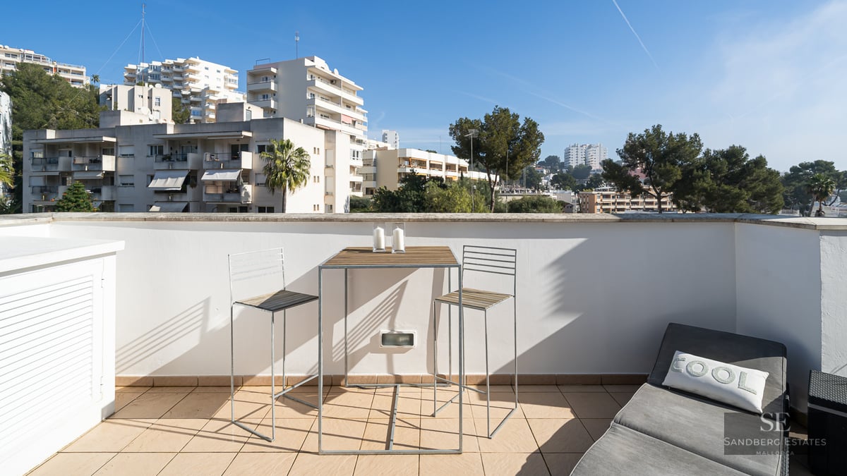 A sunlit terrace with a high-top wooden table, two bar chairs, and a grey sun lounger overlooking city buildings.