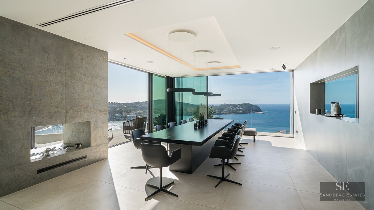 A modern dining room with a long black table, stone fireplace, and floor-to-ceiling windows overlooking the ocean.