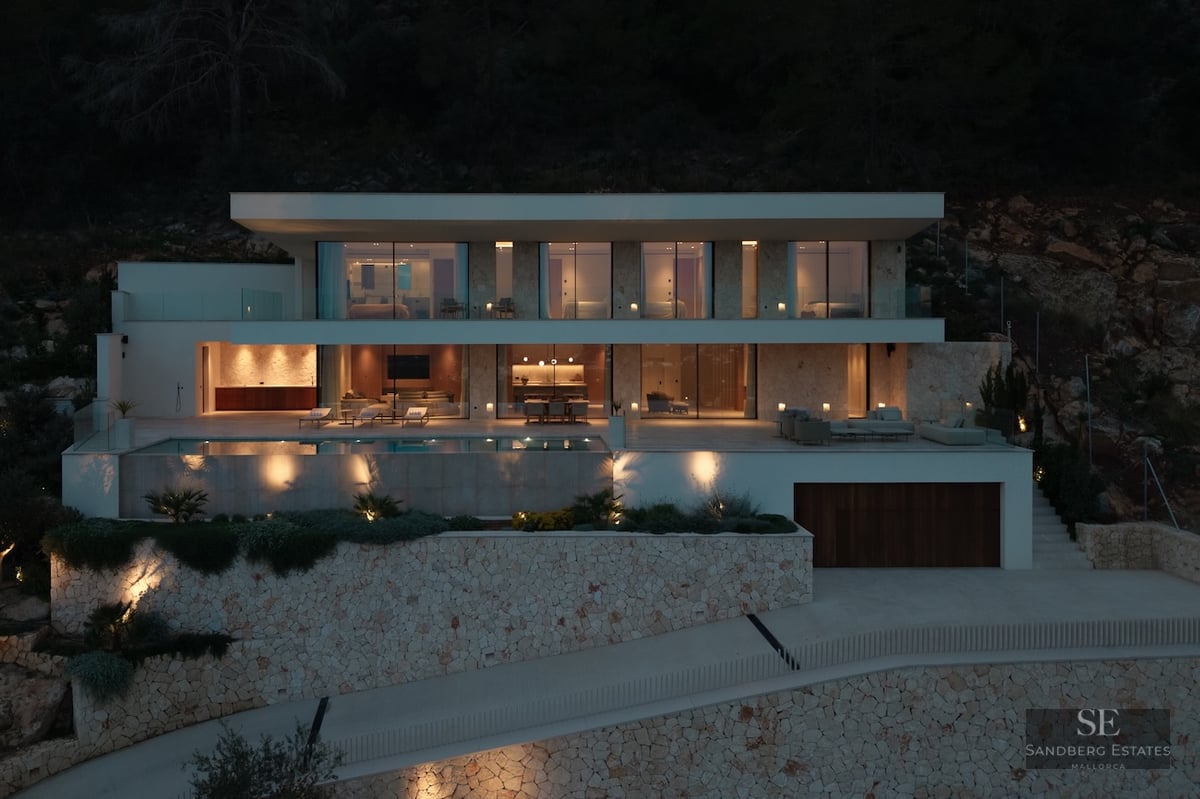 Illuminated nighttime facade of a three-level modern villa featuring an infinity pool and natural stone walls.
