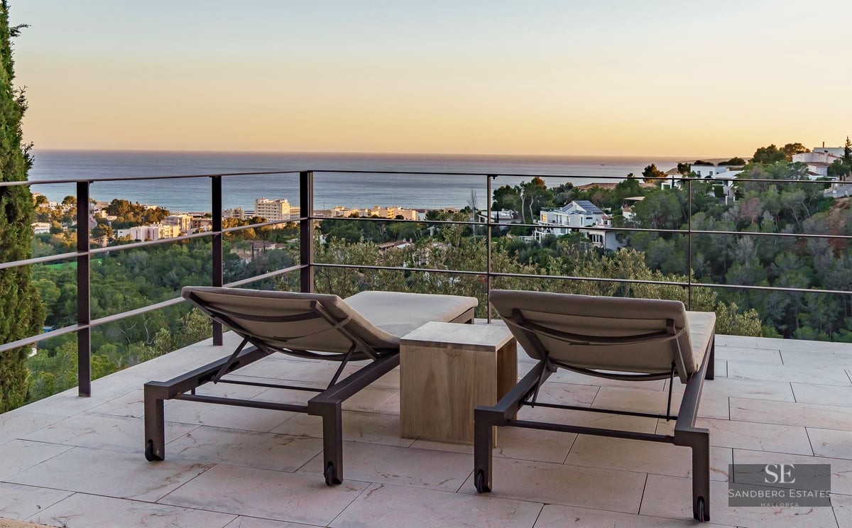 Two modern sun loungers on a stone terrace facing a metal railing with panoramic sea and coastal views at sunset.