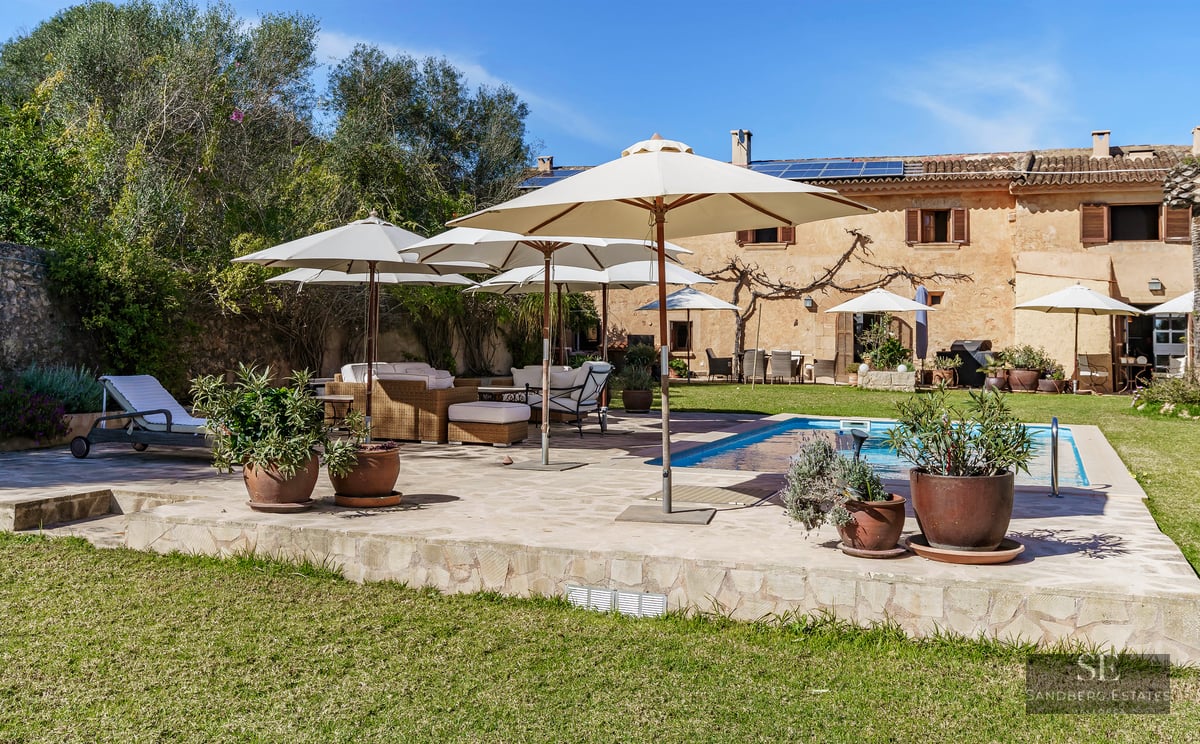 Large blue swimming pool surrounded by a stone deck with white umbrellas and wicker lounge furniture in a sunny garden.