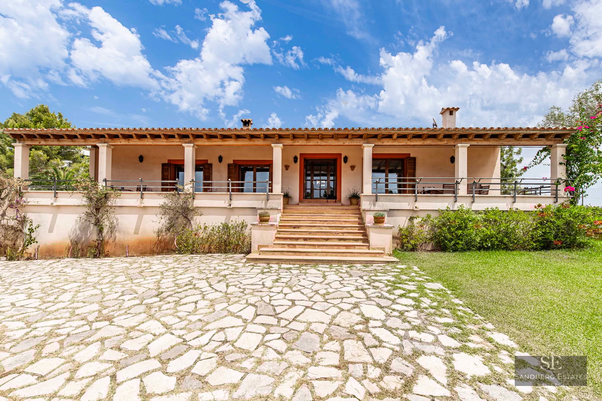 Front view of a one-story Mediterranean villa with a stone driveway, steps, and a covered terrace under a blue sky.