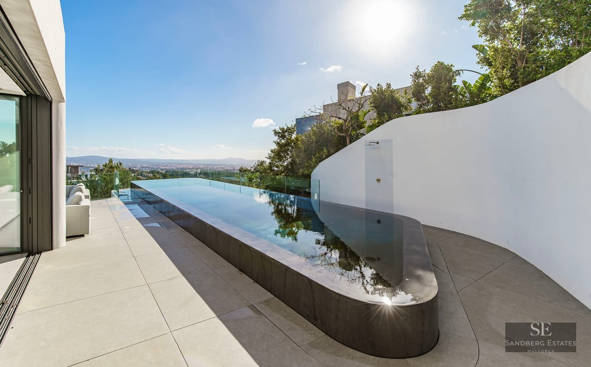 A sleek, long infinity pool on a minimalist white terrace overlooking a distant city under a bright blue sky.