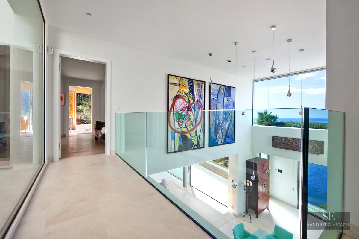 Modern hallway with glass railing, large paintings, and sea views through expansive windows.