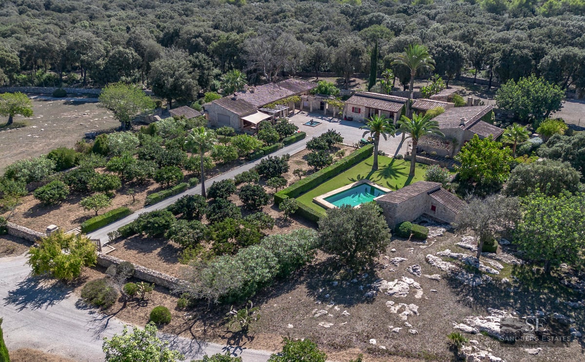 Bird's eye view of a stone villa estate with a swimming pool, palm trees, and lush gardens surrounded by forest.