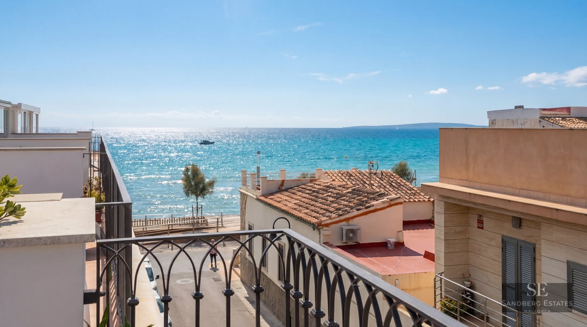High-angle view from a wrought iron balcony looking out over terracotta rooftops toward a sparkling turquoise sea.