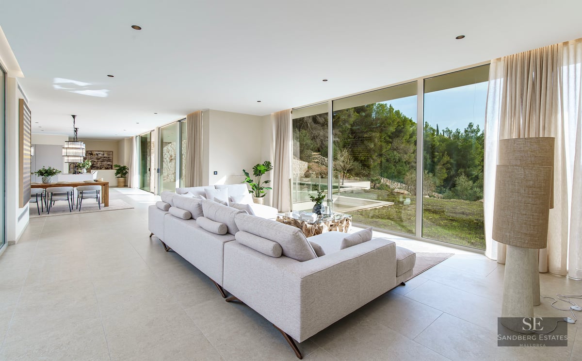 Spacious modern living room with a large grey sofa and floor-to-ceiling windows overlooking a lush green forest.