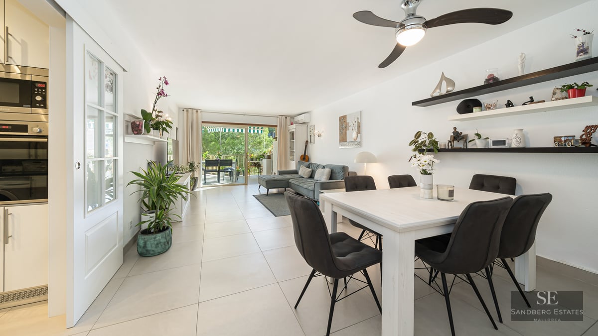 Espace de vie lumineux avec table à manger blanche, canapé gris et baies vitrées ouvrant sur un balcon verdoyant.