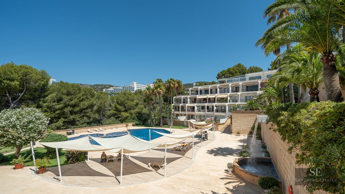 A large blue swimming pool surrounded by a stone terrace with white shade sails and palm trees under a clear blue sky.