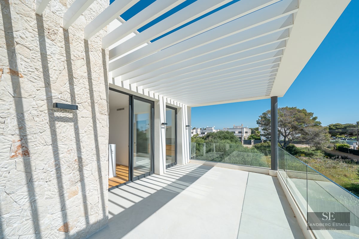 Bright outdoor terrace with a white slatted pergola casting shadows on a textured stone wall and glass railing.
