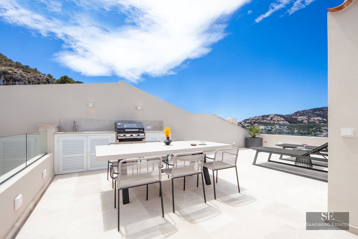 Modern rooftop terrace featuring an outdoor kitchen, dining table, and sun loungers with panoramic coastal views.
