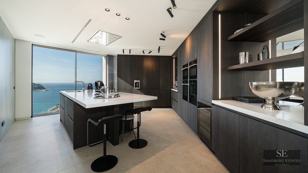 Contemporary dark wood kitchen with a large white island and panoramic ocean views through floor-to-ceiling windows.