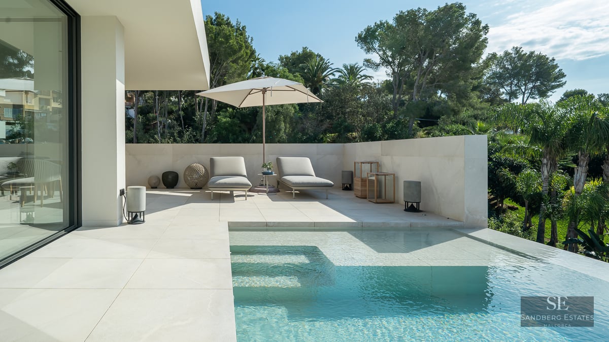 Modern luxury infinity pool with white stone deck, sun loungers, umbrella, and a background of green trees.