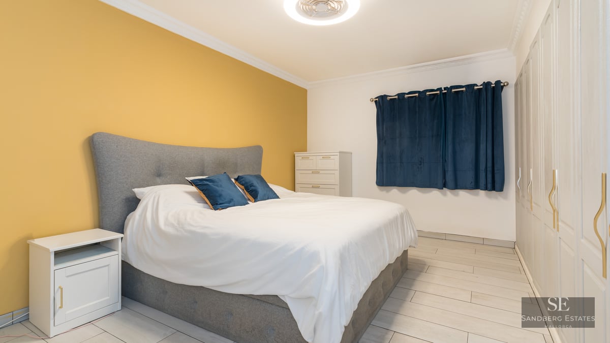 Une chambre lumineuse avec lit gris, mur jaune, placards blancs et rideaux bleu foncé.
