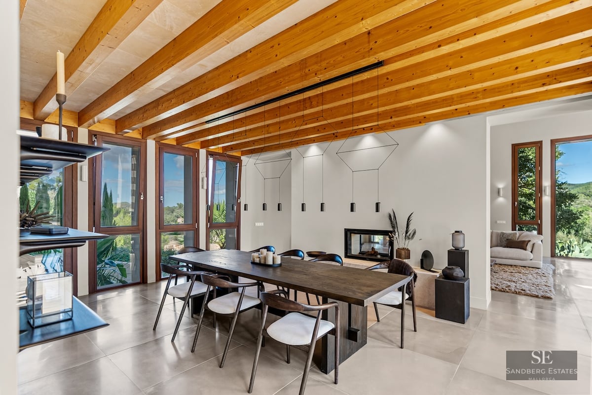 Large wooden dining table under a beamed ceiling with floor-to-ceiling windows showing nature views.